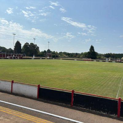 A sunny view of an empty football pitch with well-maintained green grass, surrounded by low red and white barriers. In the background, there are small buildings, trees, and tall floodlight poles under a blue sky with scattered clouds.