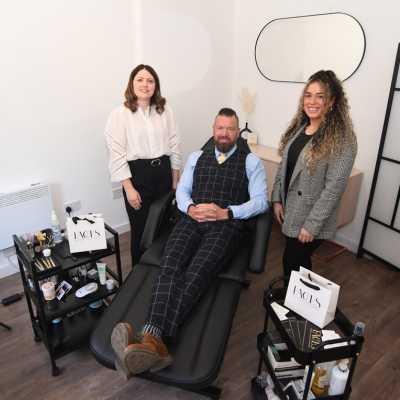 Three people in a salon, one person sat in a black beauty chair smiling