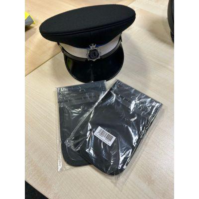 A black police cap with a silver band and badge, next to two sealed black Faraday pouches on a wooden surface.