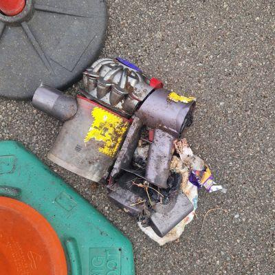 A damaged metal automotive component, covered in dirt and debris, sits on a grey surface next to coloured plastic objects.