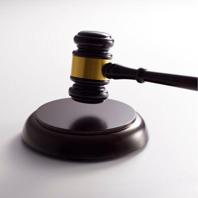 A judge’s gavel with a gold band rests on a matching dark wooden sound block. The background is plain and light-coloured, drawing attention to the gavel and sound block.