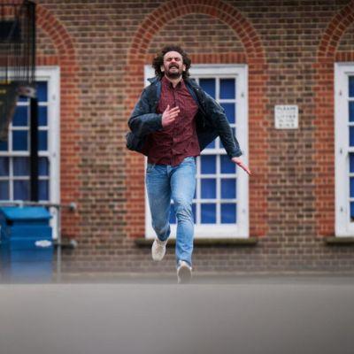 Person running outdoors on a paved surface in front of a brick building with white-framed windows, wearing a dark jacket, red shirt, and blue jeans.