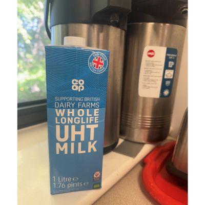 A blue Co-op brand whole long-life UHT milk carton, labelled 1 litre, sits on a kitchen counter. The carton displays a British flag emblem and white text. In the background are two stainless steel thermal carafes and part of a red tray.
