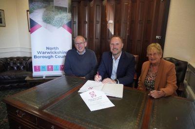 Three people sitting at a table with documents. The person in the middle is signing a paper. A banner behind them reads &#039;North Warwickshire Borough Council&#039; with the word &#039;THRIVE&#039; underneath.