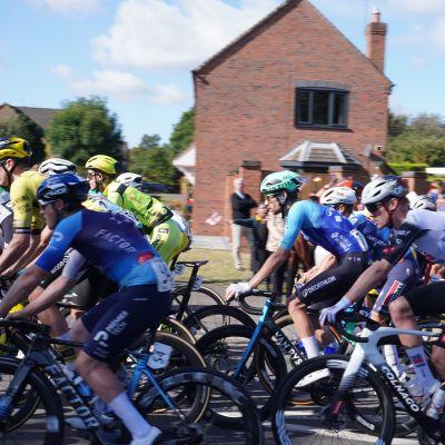 A group of cyclists in bright jerseys is competing in a race, with a brick house in the background under a clear blue sky.